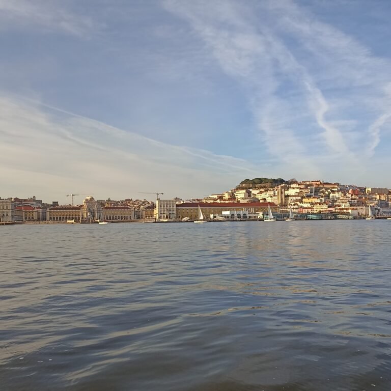 View from a sailboat deck with the 25 de Abril Bridge in the distance at sunset.