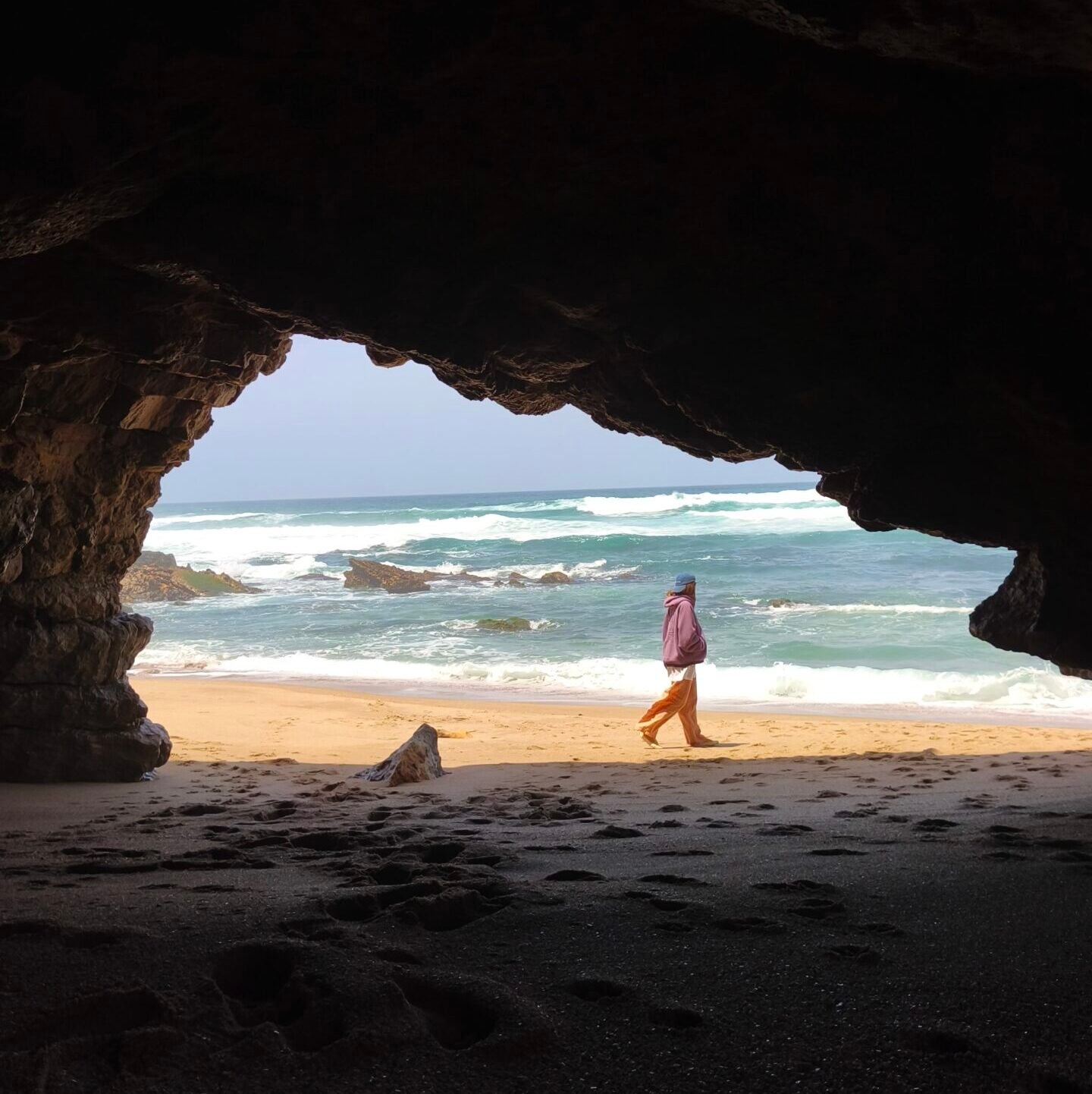 A person walking on the sand of a deserted beach, viewed from inside a coastal cave in Cascais.
