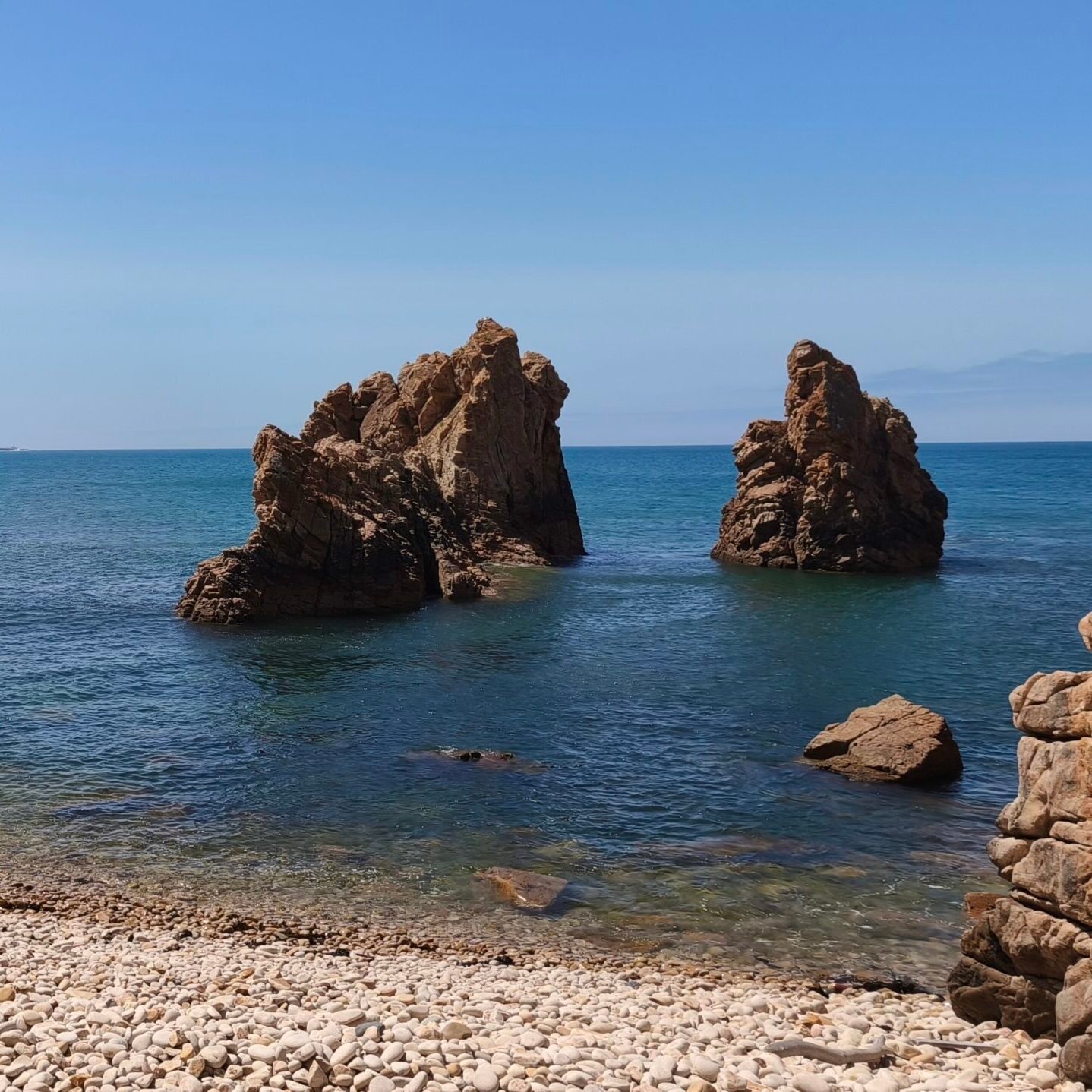 Large rock formations emerging from a calm sea, viewed from an isolated pebble beach in Cascais.